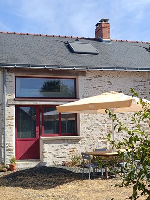 Una casa de piedra con puerta y ventanas rojas, una chimenea y un patio con mesa y silla.