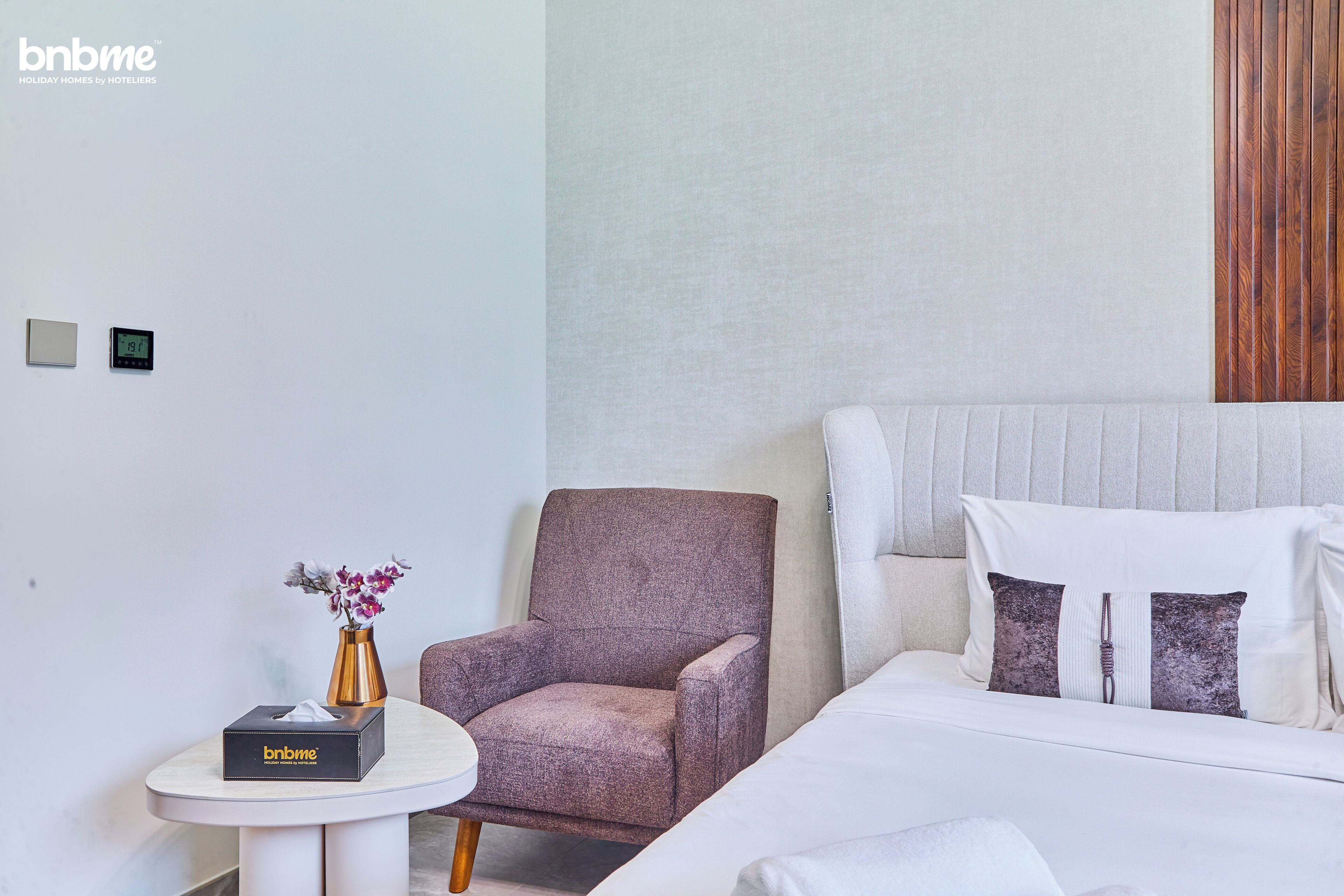 Ein Hotelzimmer mit einem Bett, einem Sessel, einem kleinen Tisch und einem an der Wand montierten Thermostat.