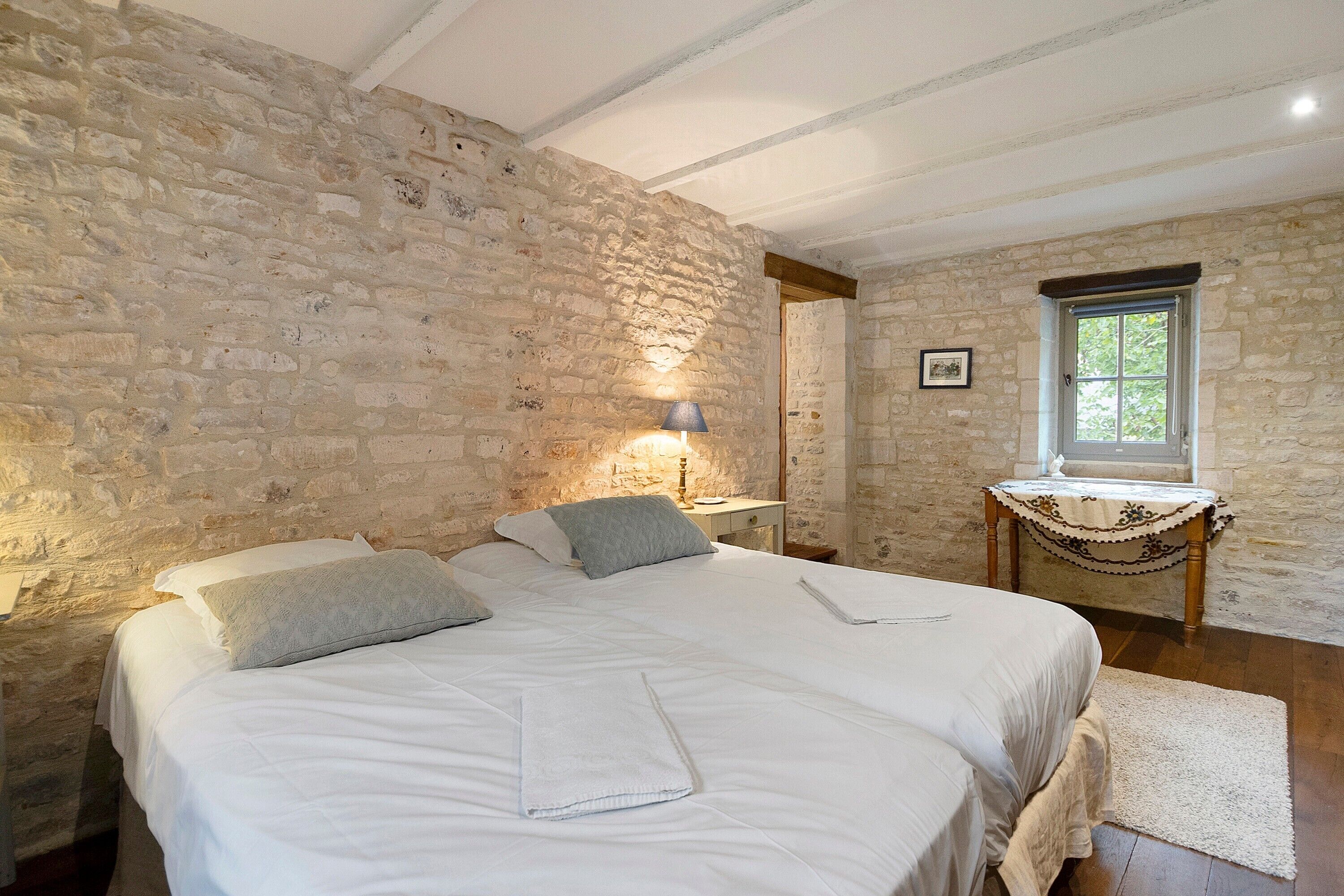 Ein Schlafzimmer mit Stehwänden, einem Bett mit weißer Bettwäsche, einem Nachttisch, einer Lampe und einem Fenster mit Blick auf Grünflächen.