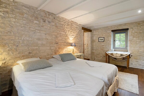 Une chambre avec un mur en pierre, un lit avec une literie blanche, une table de chevet, une lampe et une fenêtre donnant sur un paysage verdoyant.