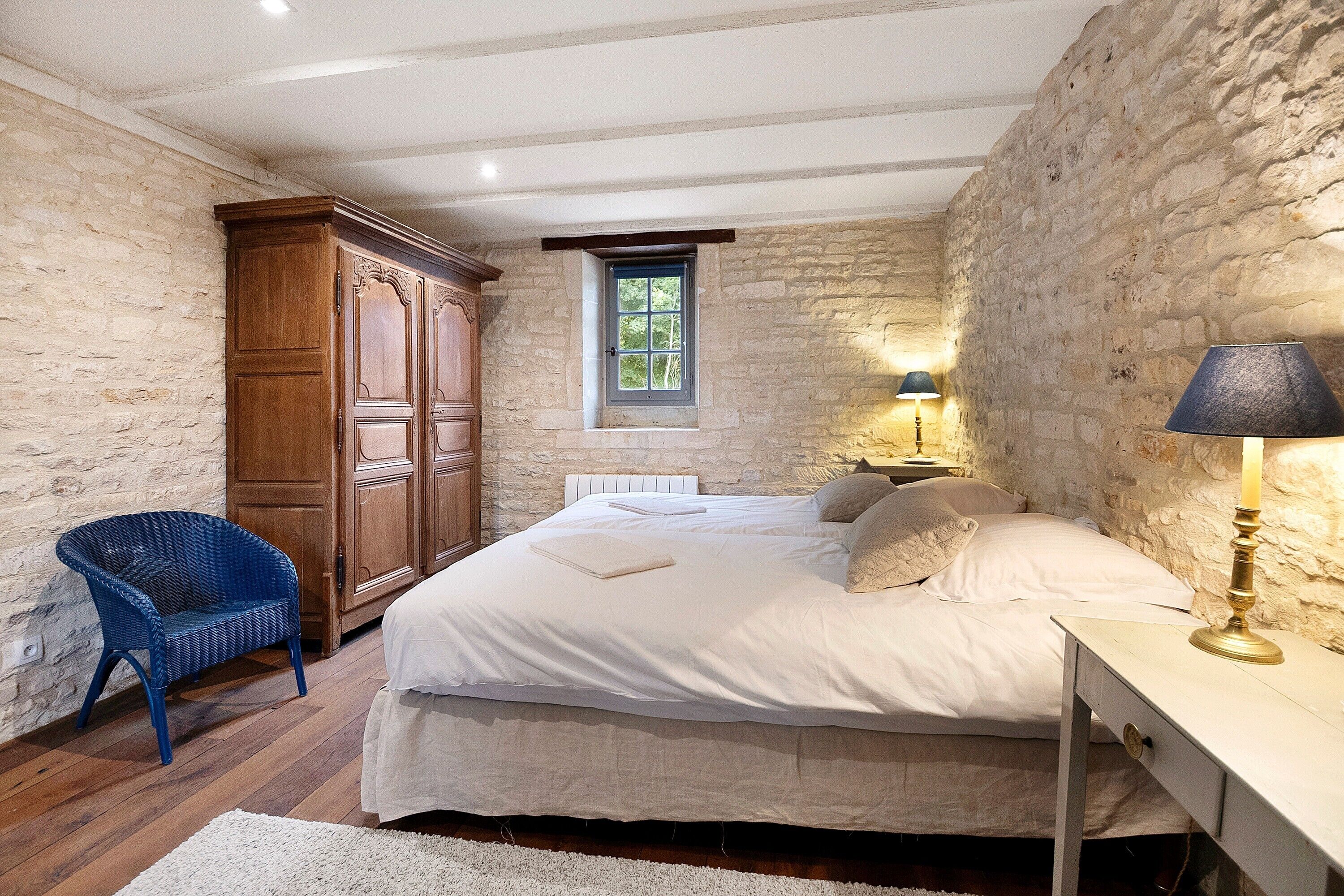 A bedroom with a stone wall, a wooden wardrobe, a bed with white linens, a blue chair, and a bedside table with a lamp.