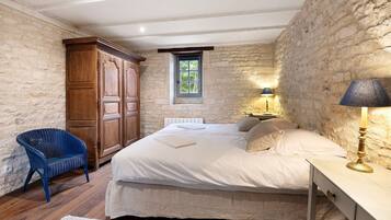 A bedroom with a stone wall, a wooden wardrobe, a bed with white linens, a blue chair, and a bedside table with a lamp.