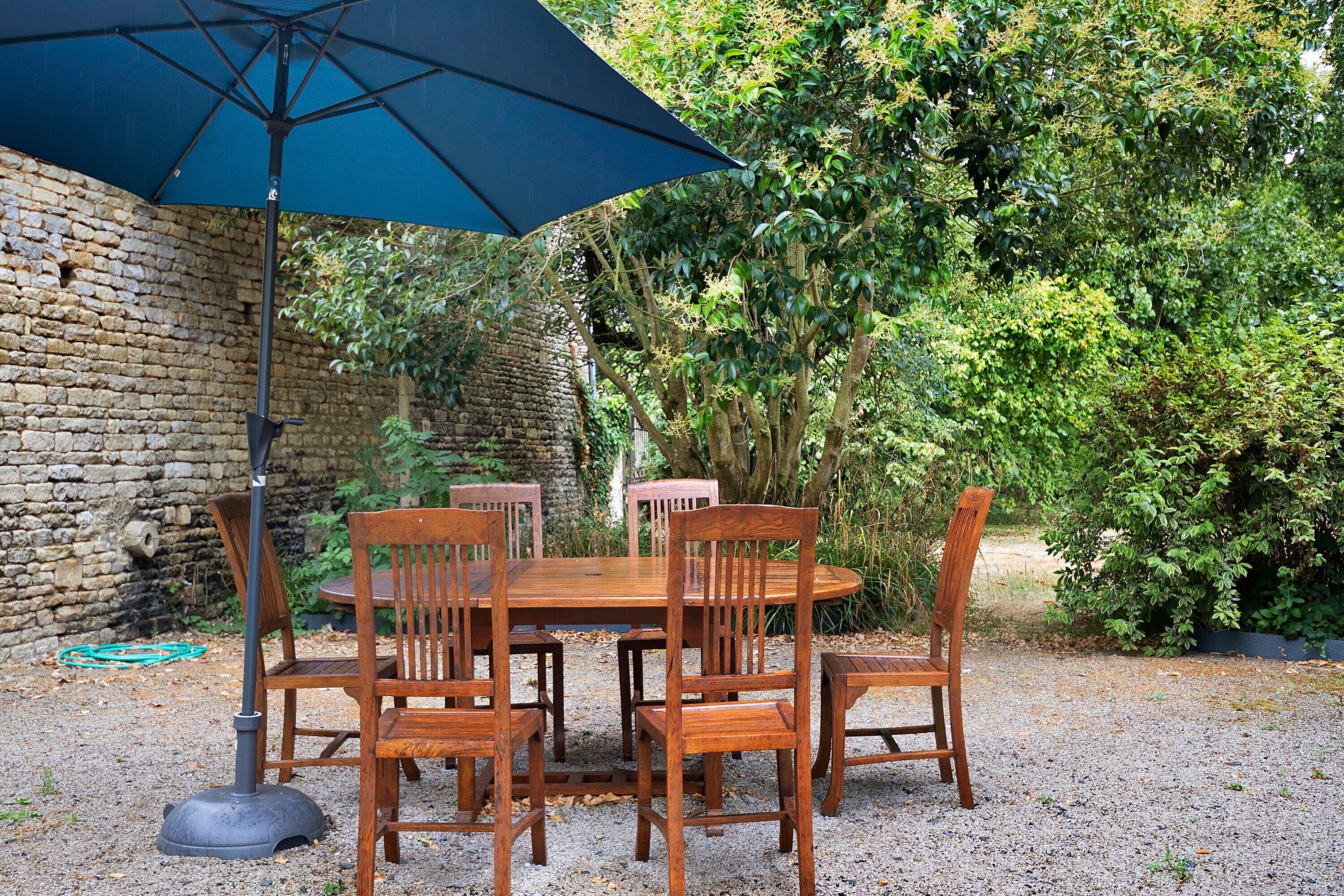 Une table et des chaises en bois sont disposées à l’extérieur, sous un parasol bleu.