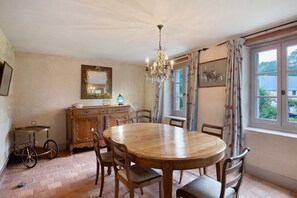 Uma sala de jantar com mesa e cadeiras de madeira, um candelabro, um espelho e uma janela com cortinas.