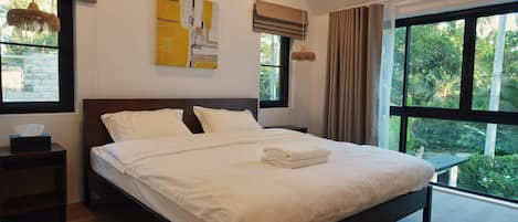A modern bedroom with a large bed, a bedside table, a window with curtains, and a view of greenery outside.