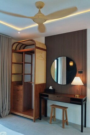 Une pièce avec un ventilateur de plafond, une armoire en bois dotée d’une porte en osier, un miroir rond, un bureau avec une lampe et un tabouret.