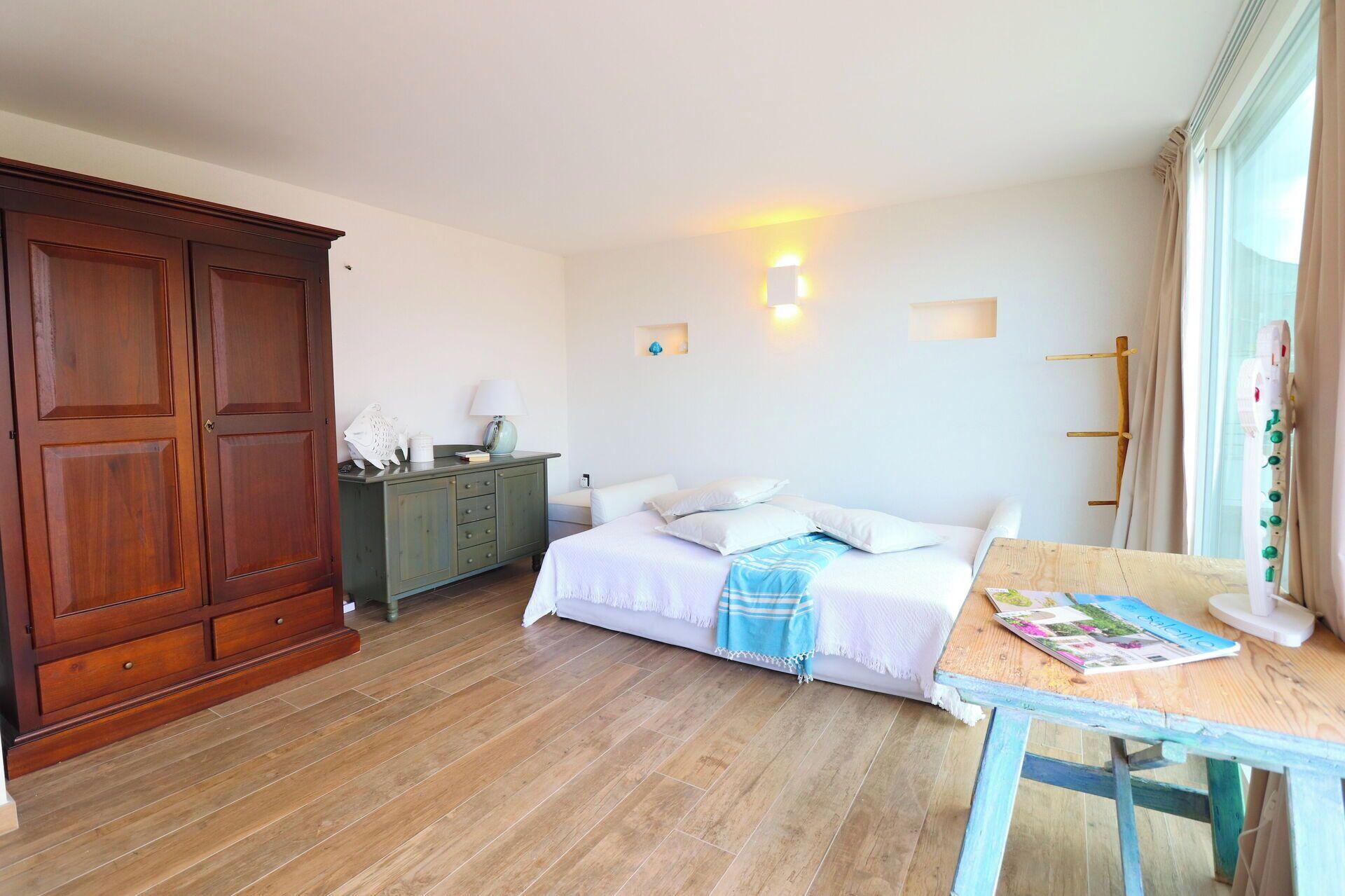 Un dormitorio con cama, un armario de madera, una mesita de noche con lámpara y una ventana con cortinas.