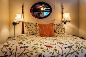A bed with a floral-patterned bedspread, two wall-mounted lamps, and a round mirror above the bed. - King Studio + Kitchenette | Oak Hills + 30 Min to Coast | Room With A View (Paso Robles)