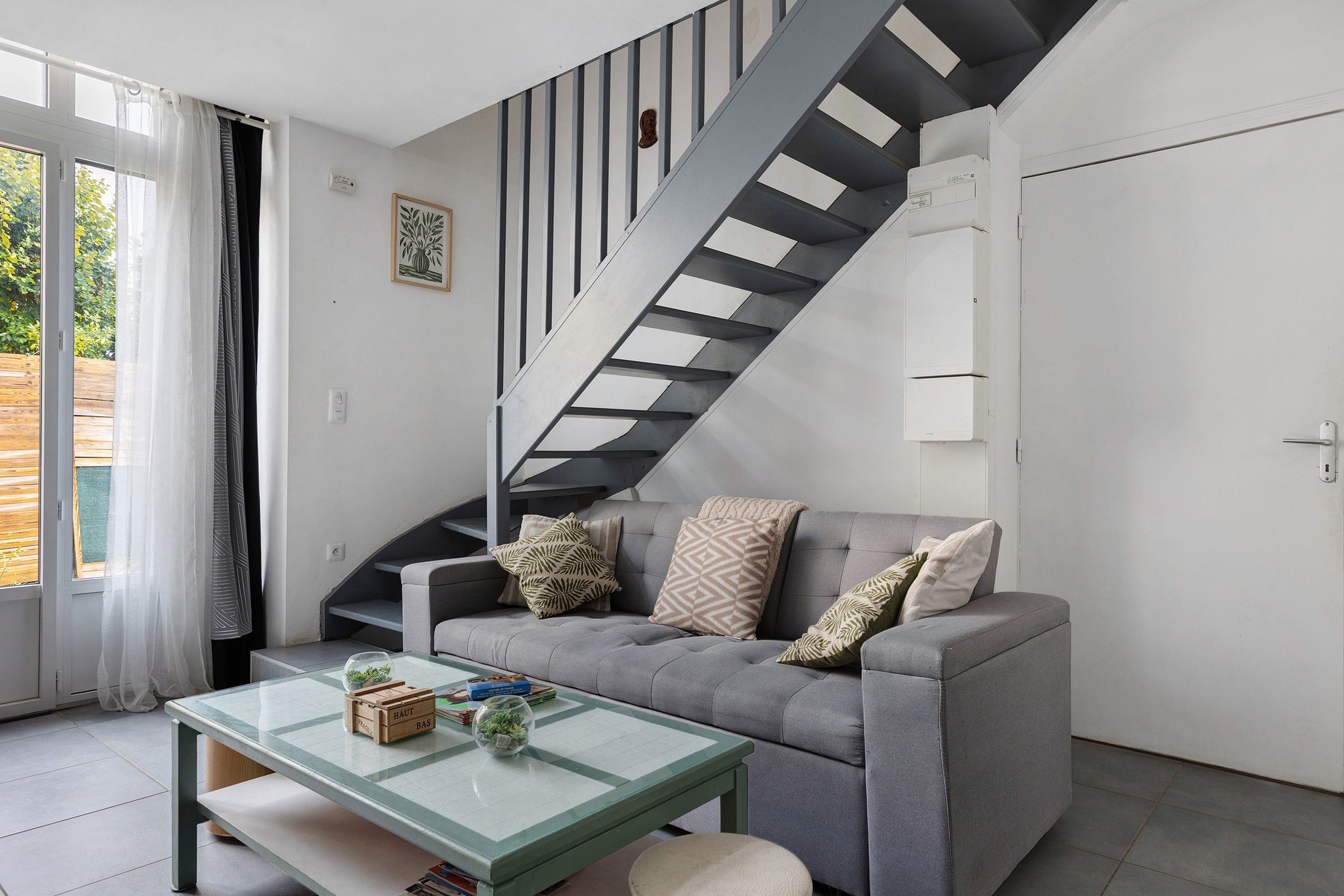 A modern living room with a grey sofa, a glass coffee table, and a staircase with black and white stripes.