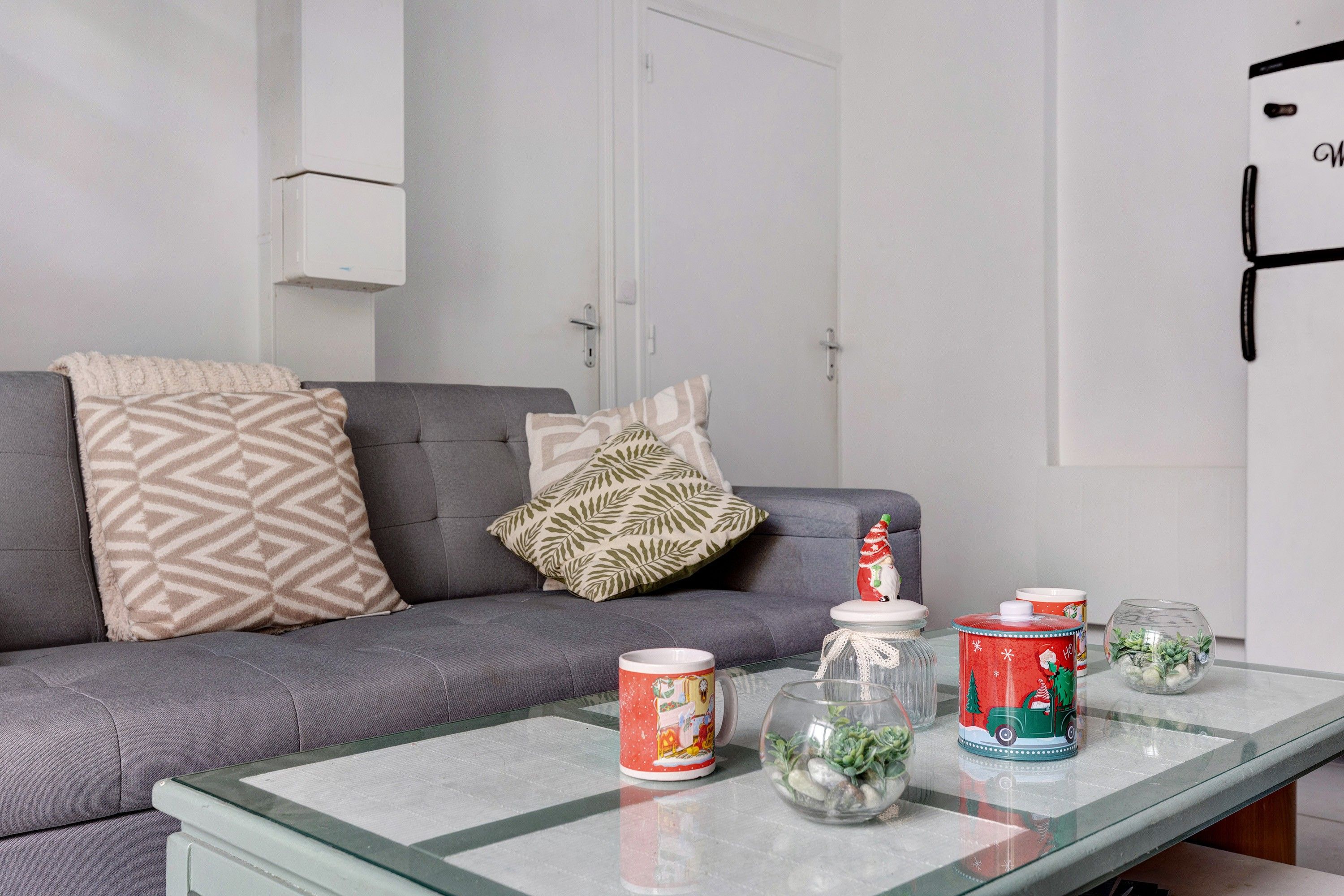 Ein Wohnzimmer mit einem grauen Sofa, gemusterten Kissen, einem Glastisch und dekorativen Gegenständen, darunter eine Tasse, ein Glasbehälter mit Pflanzen und eine weihnachtliche Dose.