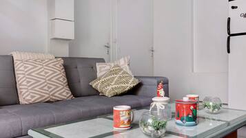 Ein Wohnzimmer mit einem grauen Sofa, gemusterten Kissen, einem Glastisch und dekorativen Gegenständen, darunter eine Tasse, ein Glasbehälter mit Pflanzen und eine weihnachtliche Dose.