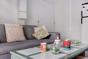 A living room with a grey sofa, patterned cushions, a glass coffee table with decorative items including a mug, a glass jar with plants, and a tin with a Christmas theme.