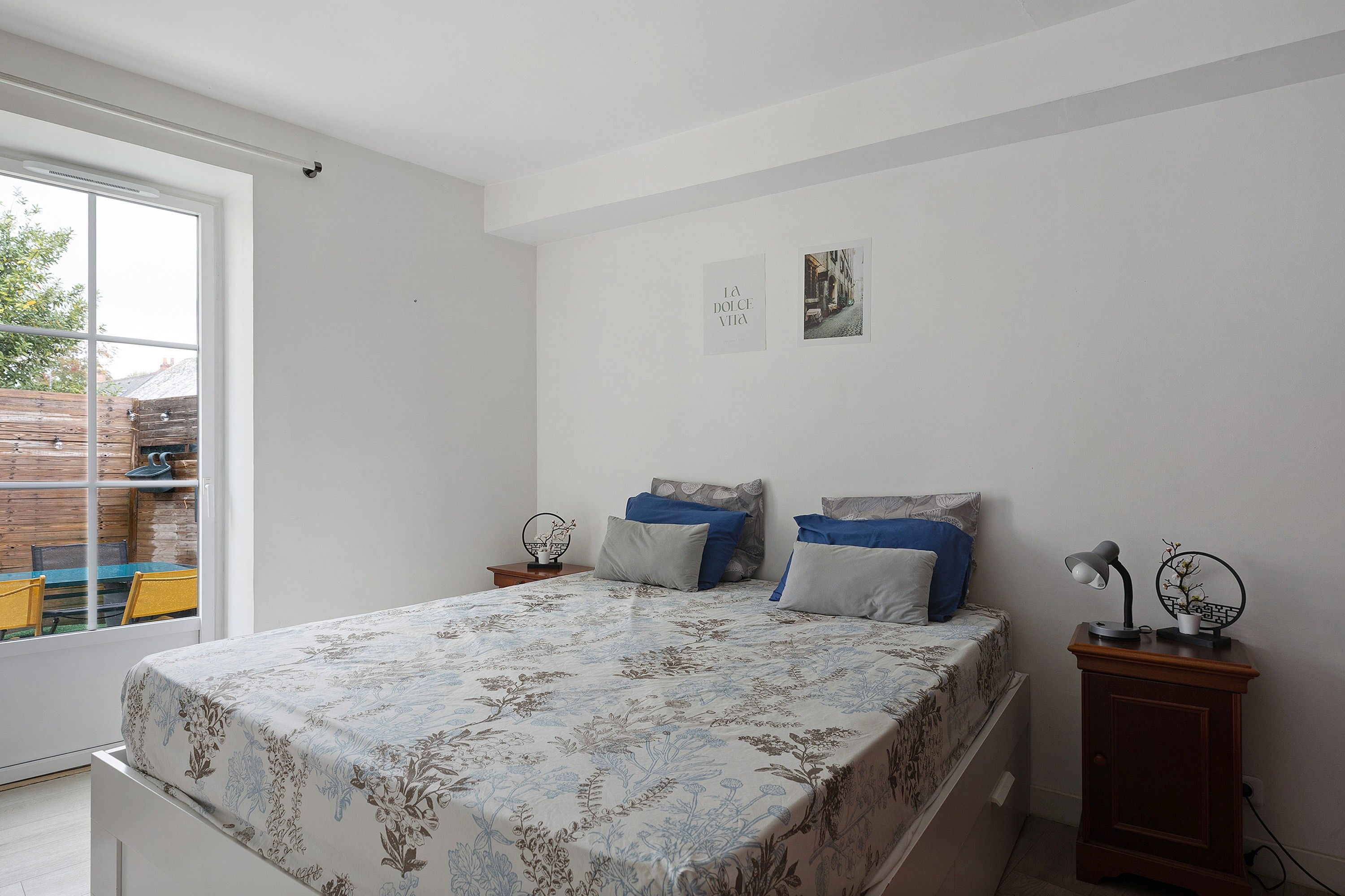 A bedroom with a bed, two bedside tables, a nightstand, a window with a view of the outside, and a wall-mounted picture.