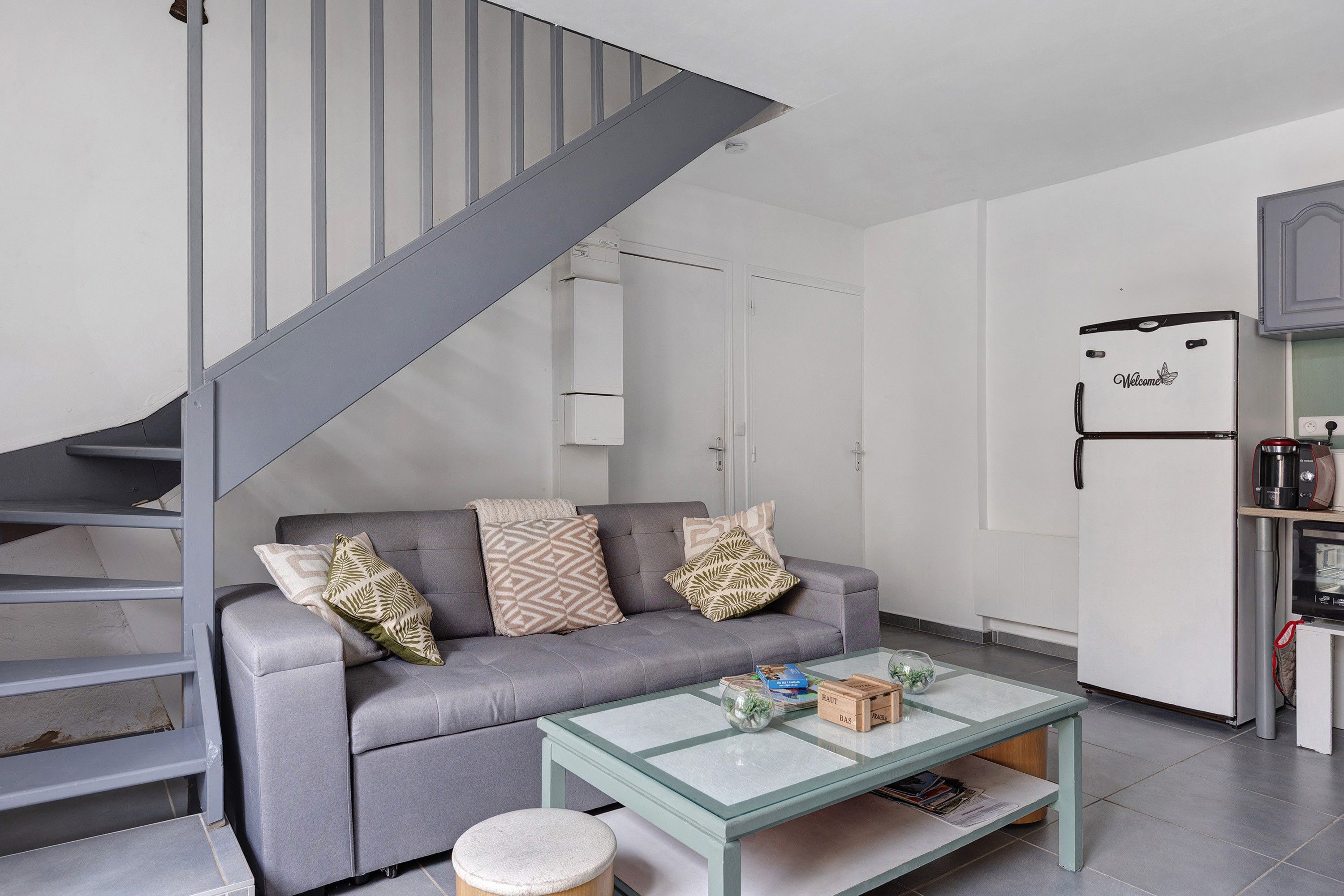 A modern living space with a grey sofa, a glass coffee table, and a kitchen area with a white refrigerator.