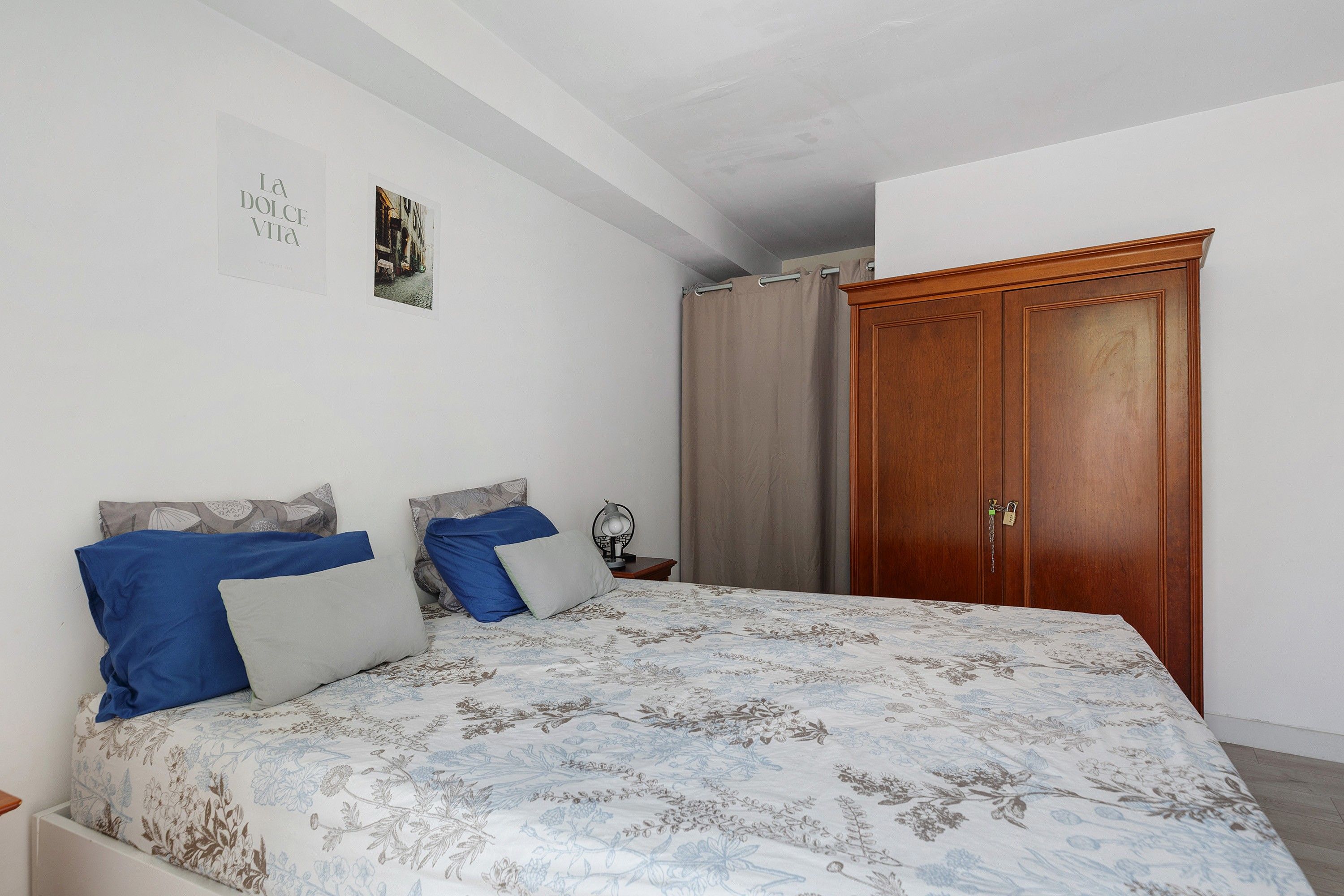 Ein Schlafzimmer mit einem Bett, einem Nachttisch, einer Lampe, einem Holzschrank und einer Wand mit einem gerahmten Bild.
