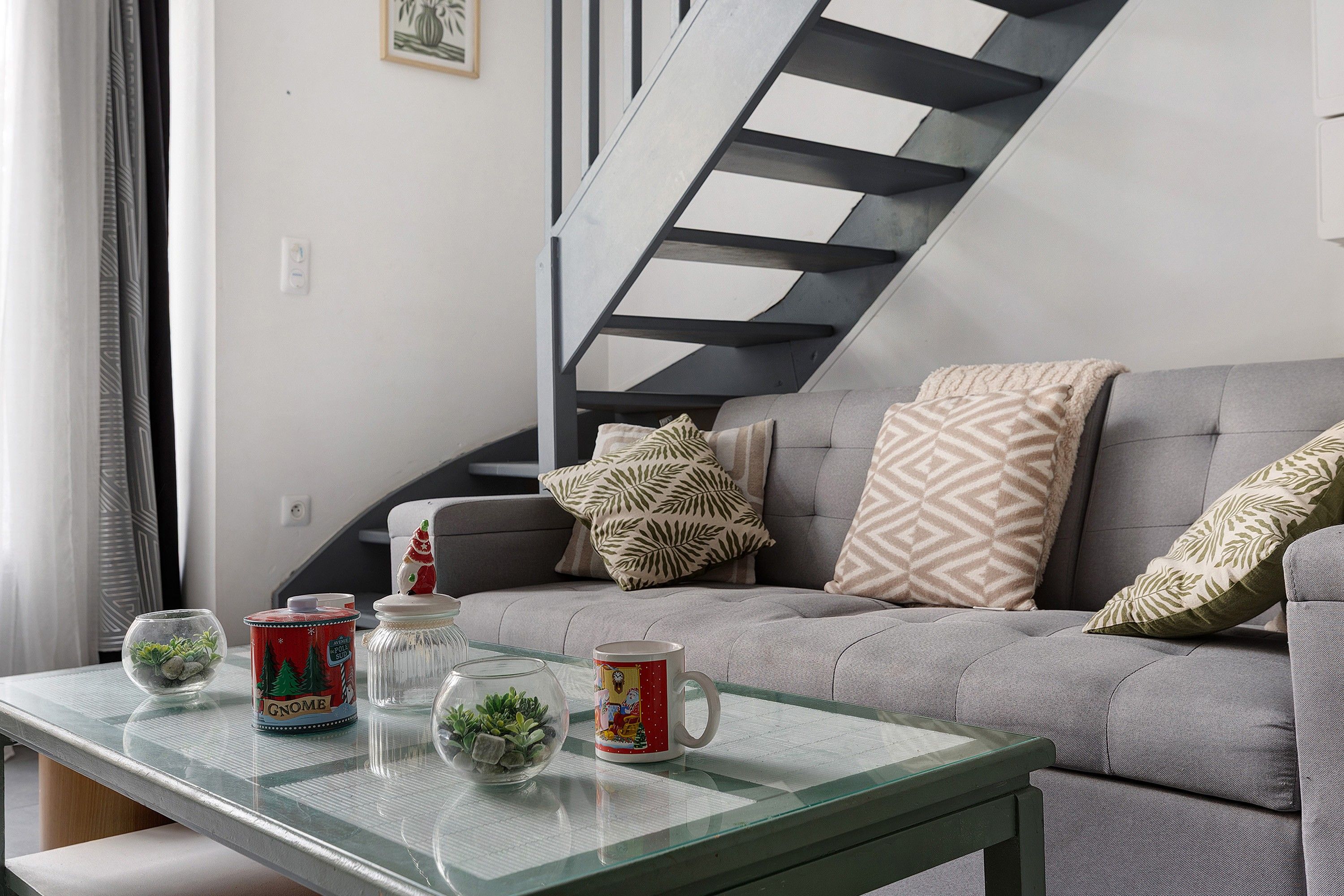 Un salon moderne avec une table basse en verre, un canapé agrémenté de coussins à motifs et un escalier au design géométrique.