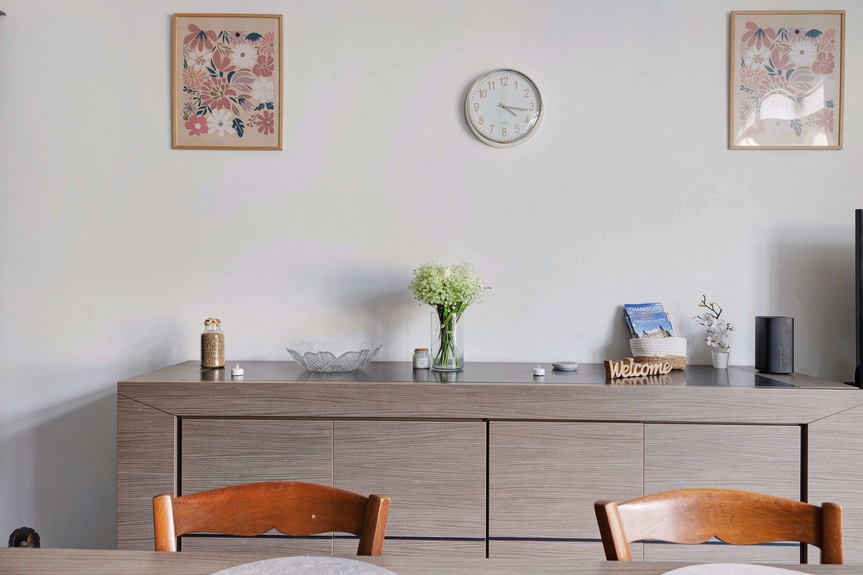 A modern dining area with a wooden table, two chairs, a wall clock, and decorative items including a vase with flowers and a framed picture.