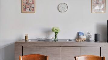 A modern dining area with a wooden table, two chairs, a wall clock, and decorative items including a vase with flowers and a framed picture.