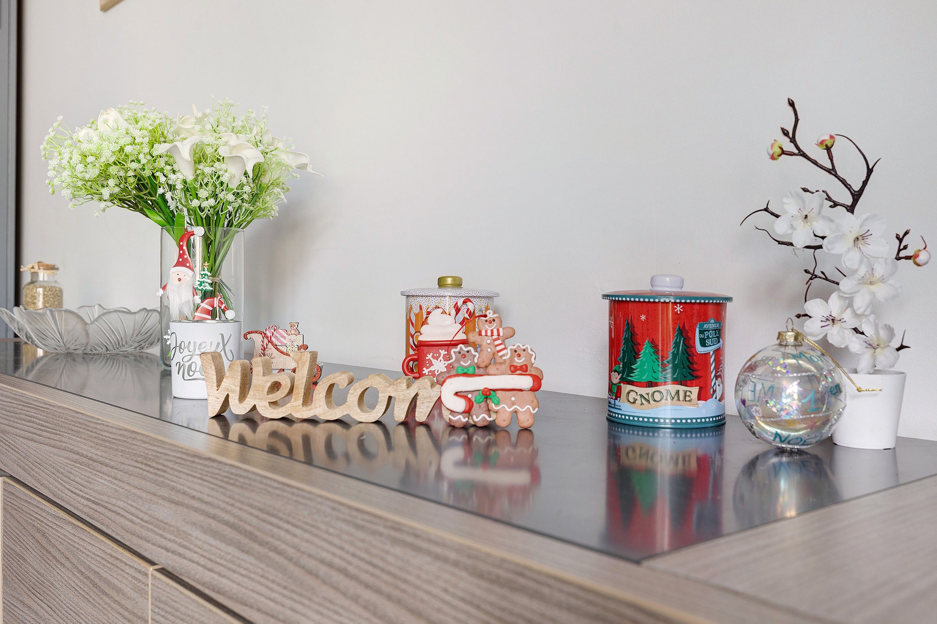 Une table ornée d’une pancarte « Bienvenue », d’un vase rempli de fleurs, d’un récipient décoré aux motifs de Noël et d’un ornement de verre.