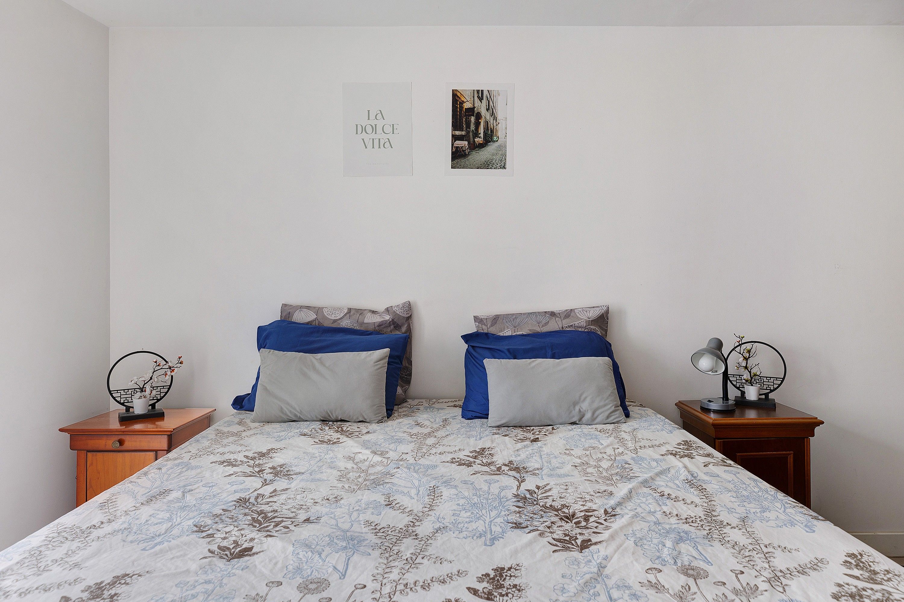 A bedroom with a bed, two nightstands, a wall-mounted shelf, and two framed pictures.