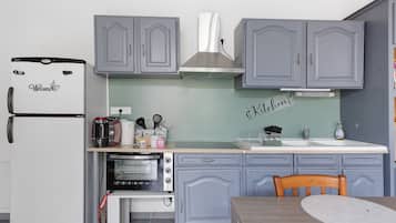 A compact kitchen with a refrigerator, toaster, kettle, and a small oven. There is a sink with a faucet, a wooden table, and a chair. The cabinets are light gray with decorative handles.