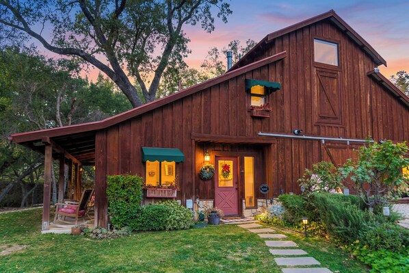 A rustic barn-style building with a porch, a front door with a wreath, and a small garden area.