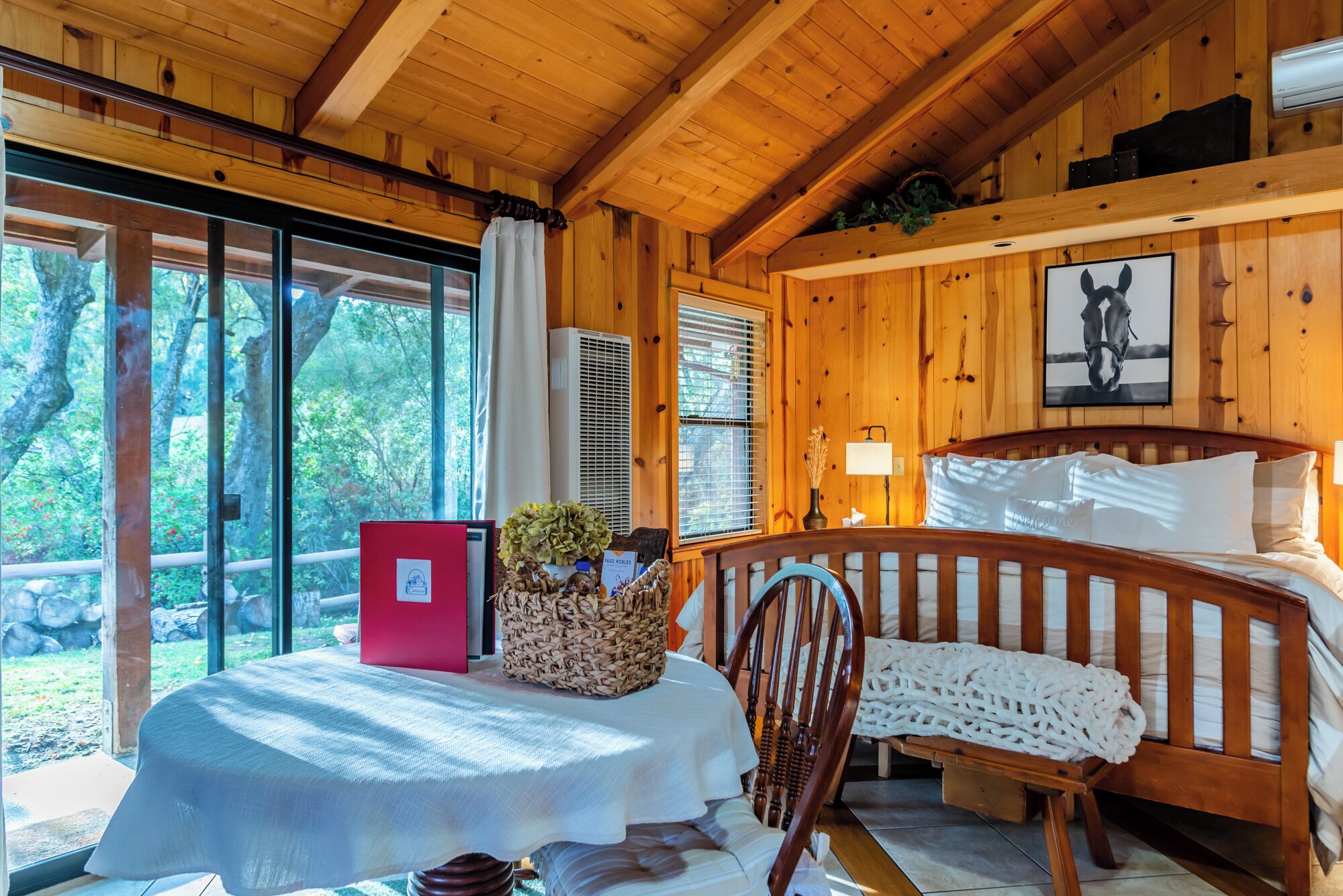 Ein Interieur einer Holzhütte mit einem Bett, einem Tisch mit einer roten Ordner, einem Stuhl und einem großen Fenster mit Blick auf Bäume.