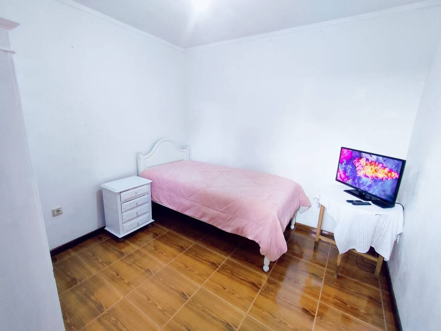 Quarto com cama, penteadeira, televisão sobre um suporte e uma mesa branca.