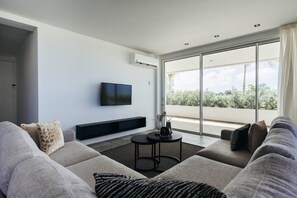 A modern living room with a large sofa, a flat-screen TV mounted on the wall, and a balcony with a view of greenery. - LUX 2BD/2Bath Condo - ONE Mambo Beach 1 by Bocobay (Willemstad)