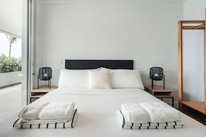 Ein modernes Hotelzimmer mit einem großen Bett, zwei Nachttischen, einem hölzernen Beistelltisch, einem schwarzen Kopfteil und einem großen Fenster mit Blick auf Grünflächen.