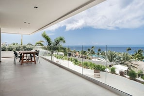 Ein geräumiger Balkon mit Holztisch und -stühlen, mit Blick auf einen Strand, an dem Palmen und das Meer zu sehen sind.