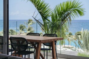 Zona pranzo all'aperto con tavolo di legno e sedie in vimini nero, con vista su una spiaggia, palme e l'oceano.