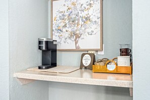 Una cafetera sobre una encimera con un cuadro enmarcado encima, una pequeña señal y una cesta con azúcar y una taza de café.