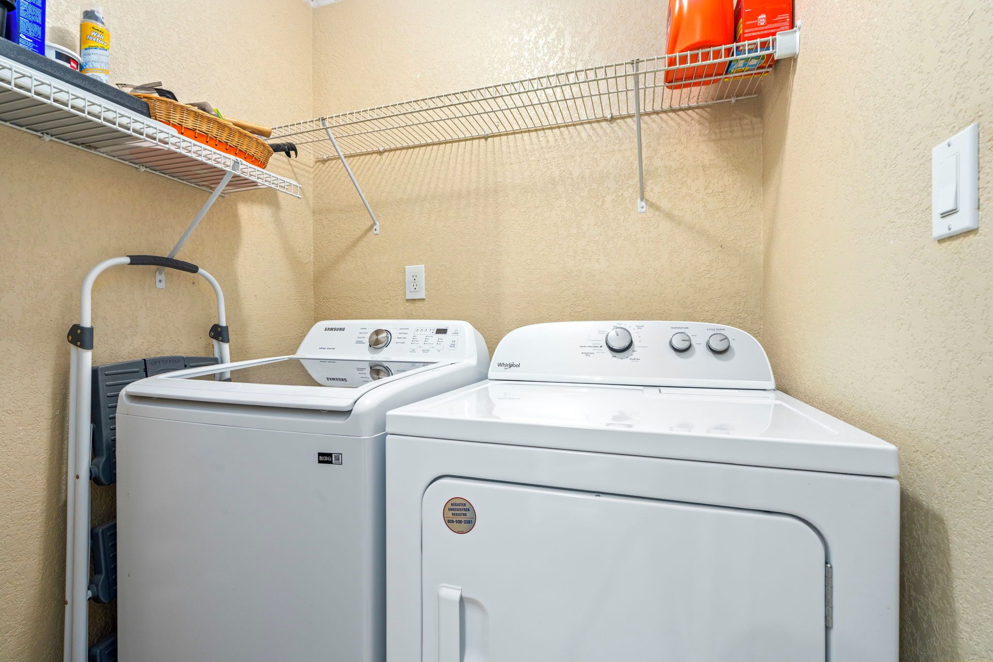 Un espace buanderie comprenant deux machines à laver et deux sèche-linge blancs, un étendoir et une étagère avec des produits de nettoyage.