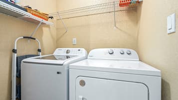 Un espace buanderie comprenant deux machines à laver et deux sèche-linge blancs, un étendoir et une étagère avec des produits de nettoyage.