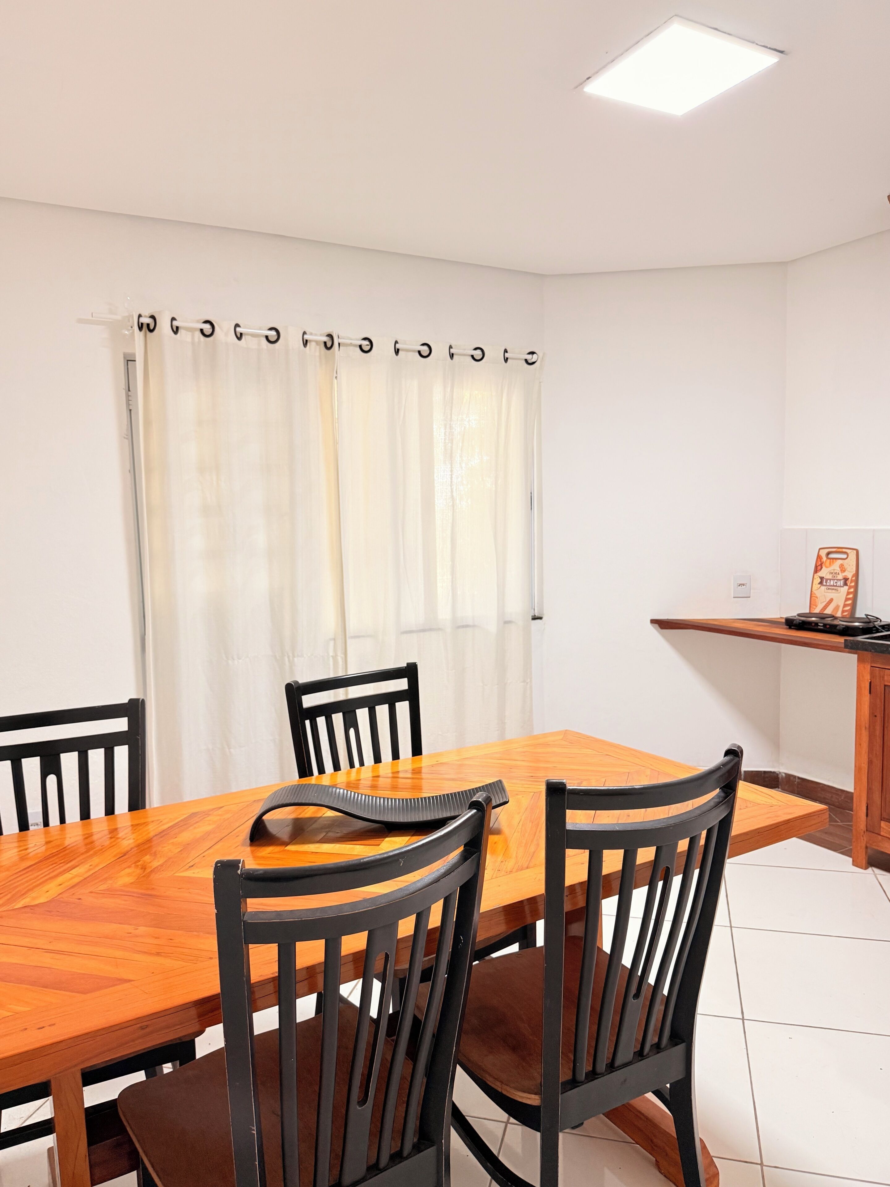 Uma área de refeições com mesa e cadeiras de madeira, uma janela com cortinas e uma pequena cozinha compacta.