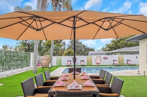 A poolside area with a table and chairs under a large umbrella, surrounded by a pool and a white wall with artwork. - Heated Pool • Jacuzzi-Fire Pit - Mini Golf - Game (Pinellas County)