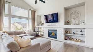 A modern living room with a sectional sofa, a flat-screen TV, a fireplace, and a large window with curtains.