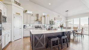 A modern kitchen with white cabinetry, a central island with stools, and a dining area with chairs. - Elevated Escape- Spacious 5BR Colorado Springs House with Hot Tub and Mountain Views (Colorado Springs)