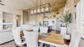 A modern kitchen with white cabinetry, a central island, and a dining area with a wooden table and white chairs. - Elevated Escape- Spacious 5BR Colorado Springs House with Hot Tub and Mountain Views (Colorado Springs)