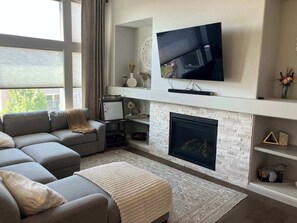 A modern living room with a sectional sofa, a flat-screen TV mounted above a fireplace, and a small side table with decorative items. - Elevated Escape- Spacious 5BR Colorado Springs House with Hot Tub and Mountain Views (Colorado Springs)