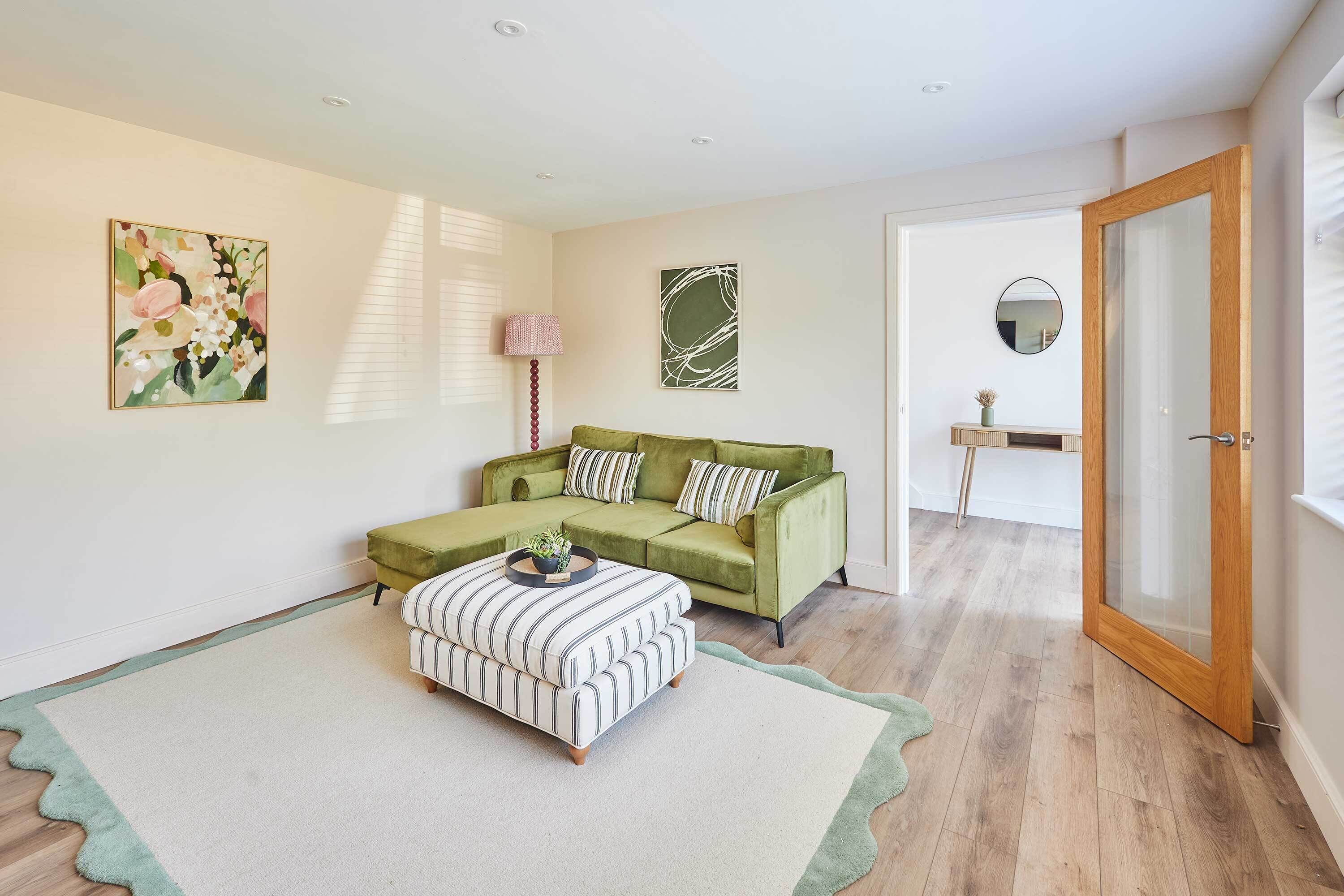 Una sala de estar con un sofá verde, un reposapiés a rayas y una alfombra verde. Hay dos cuadros en la pared y una lámpara.