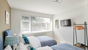 A bedroom with a bed, pillows, a bedside table, a lamp, a window with blinds, a radiator, a TV on the wall, and a clothes drying rack. - Host Stay Sandmoor (Whitby)