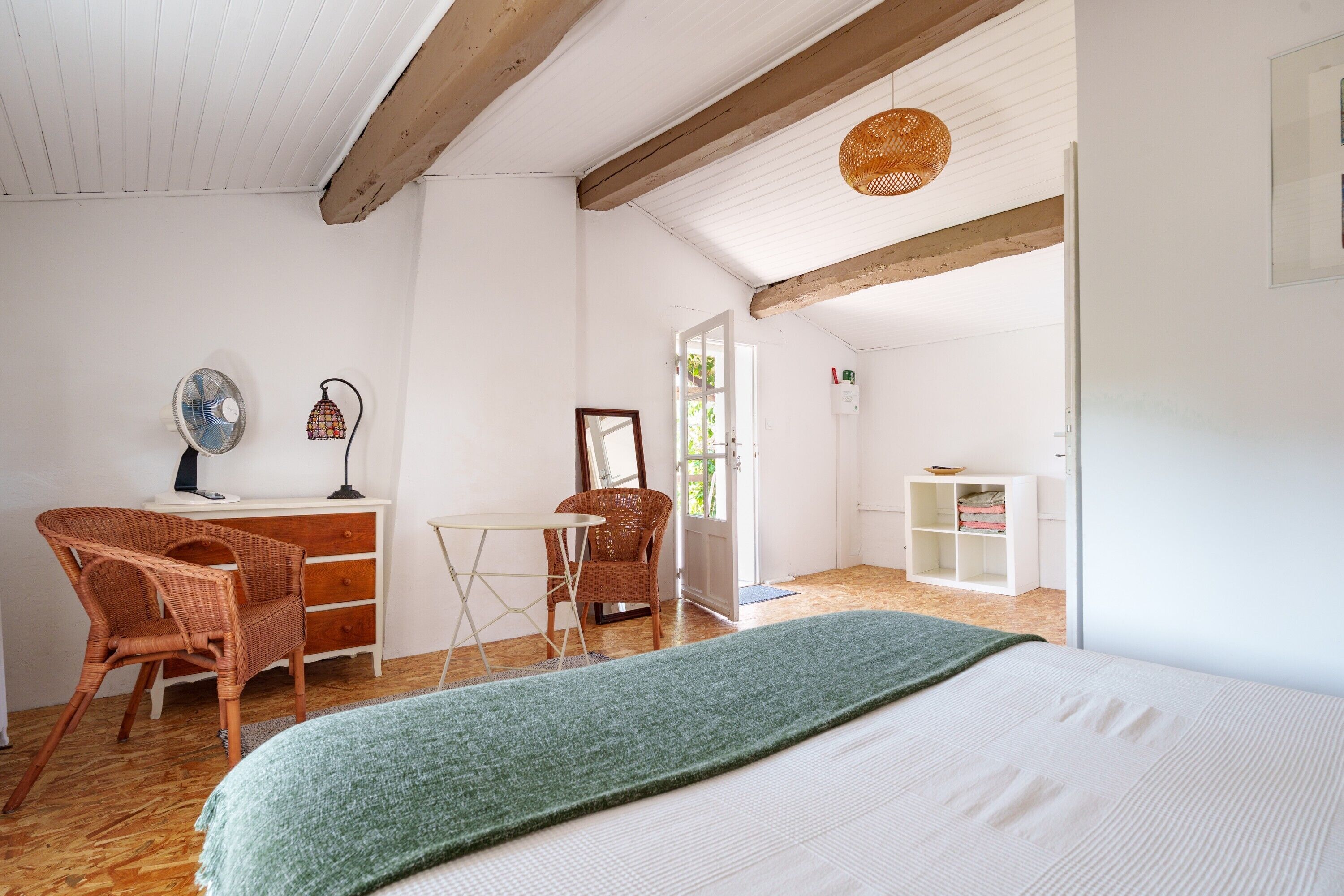 Une pièce comprenant un lit, deux fauteuils en osier, une petite table, un miroir et une étagère avec des vêtements pliés.