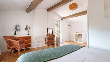 Une pièce comprenant un lit, deux fauteuils en osier, une petite table, un miroir et une étagère avec des vêtements pliés.