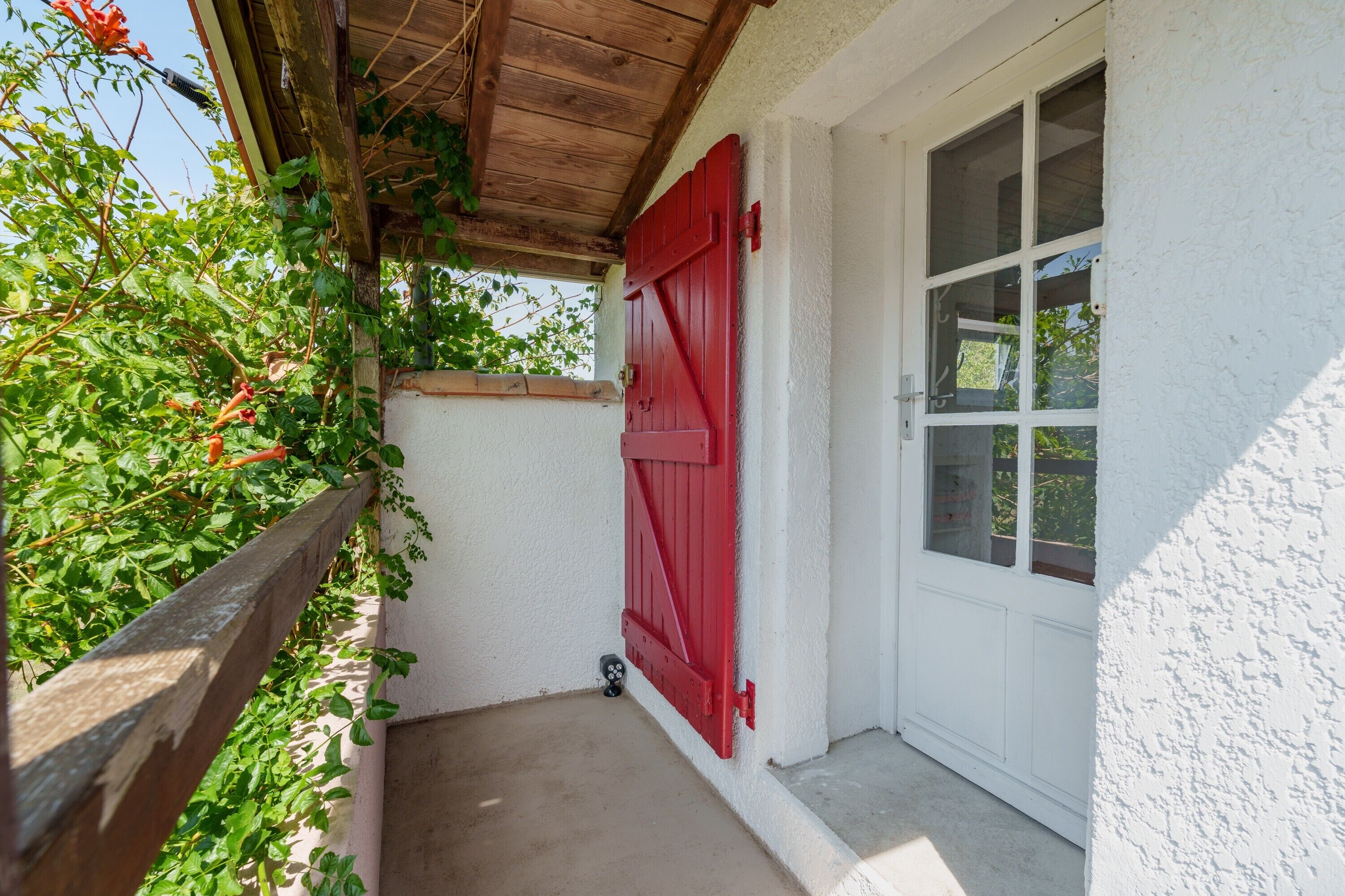 Un étroit couloir extérieur, délimité par une clôture en bois, avec une porte rouge et un mur blanc.