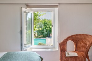 A room with a window open, a wicker chair, and a view of a swimming pool.