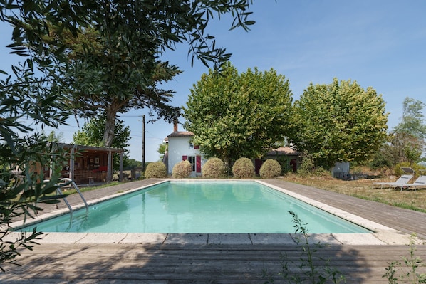 A rectangular swimming pool with a wooden deck surrounding it, a small building with a red door, and a large tree in the background.