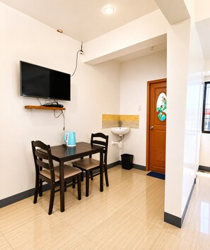 A modern dining area with a flat-screen TV mounted on the wall, a wooden dining table with four chairs, a blue kettle, and a white sink. - Metrohive Apartment 3C - Cozy Studio Near Airport & PITX (Pasay City)