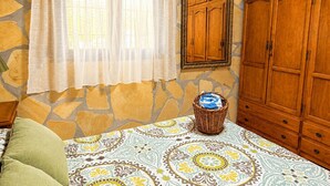 Una habitación con pared de piedra, un armario de madera, una cama con ropa de cama estampada y una ventana con cortinas traslúcidas.